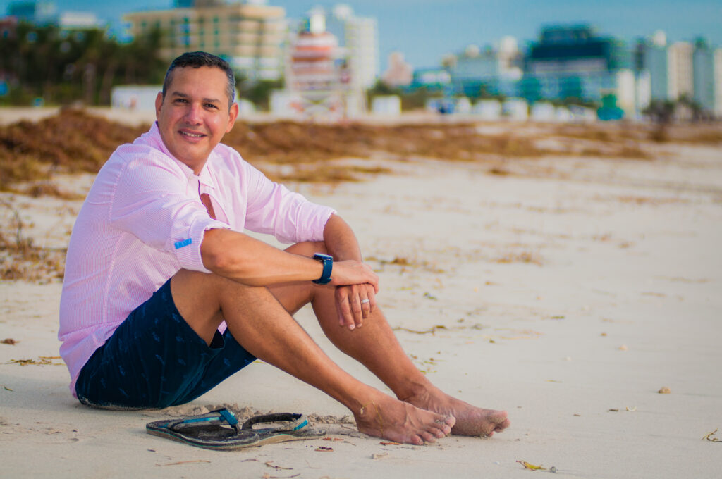 Smiling man in a pink shirt by the beach with a blurred building in the background.