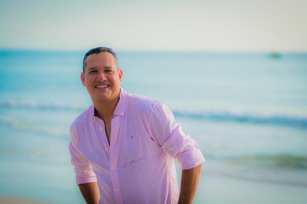 Smiling man wearing a light pink button-down shirt posing on a sunny beach, with the ocean and a soft blue sky in the background.