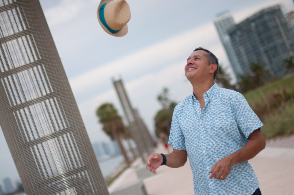 Man tossing a straw hat in the air near the waterfront with palm trees and skyline behind him.