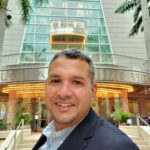 Smiling man in a blazer taking a selfie outside the Arsht Center in Miami, with palm trees and lit entrance behind him.